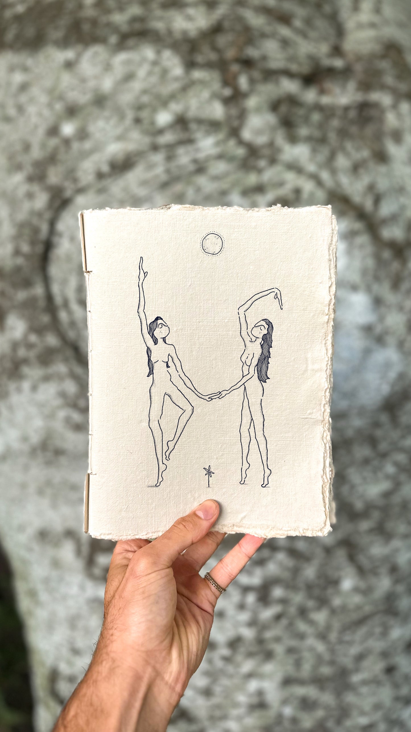 Hand holding a fineline drawing on white textured paper against a stone background. The drawing shows two dancing figures with long dark hair holding hands underneath a full moon. A tiny flower is between them.