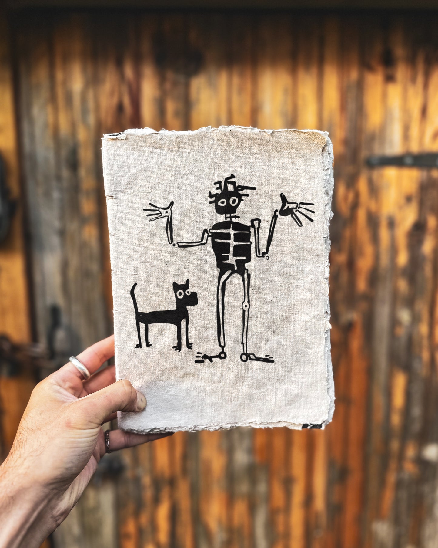 Bold drawing in black in of a skeleton shrugging and a dog beside him. The drawing is behind held in front of a wooden door.