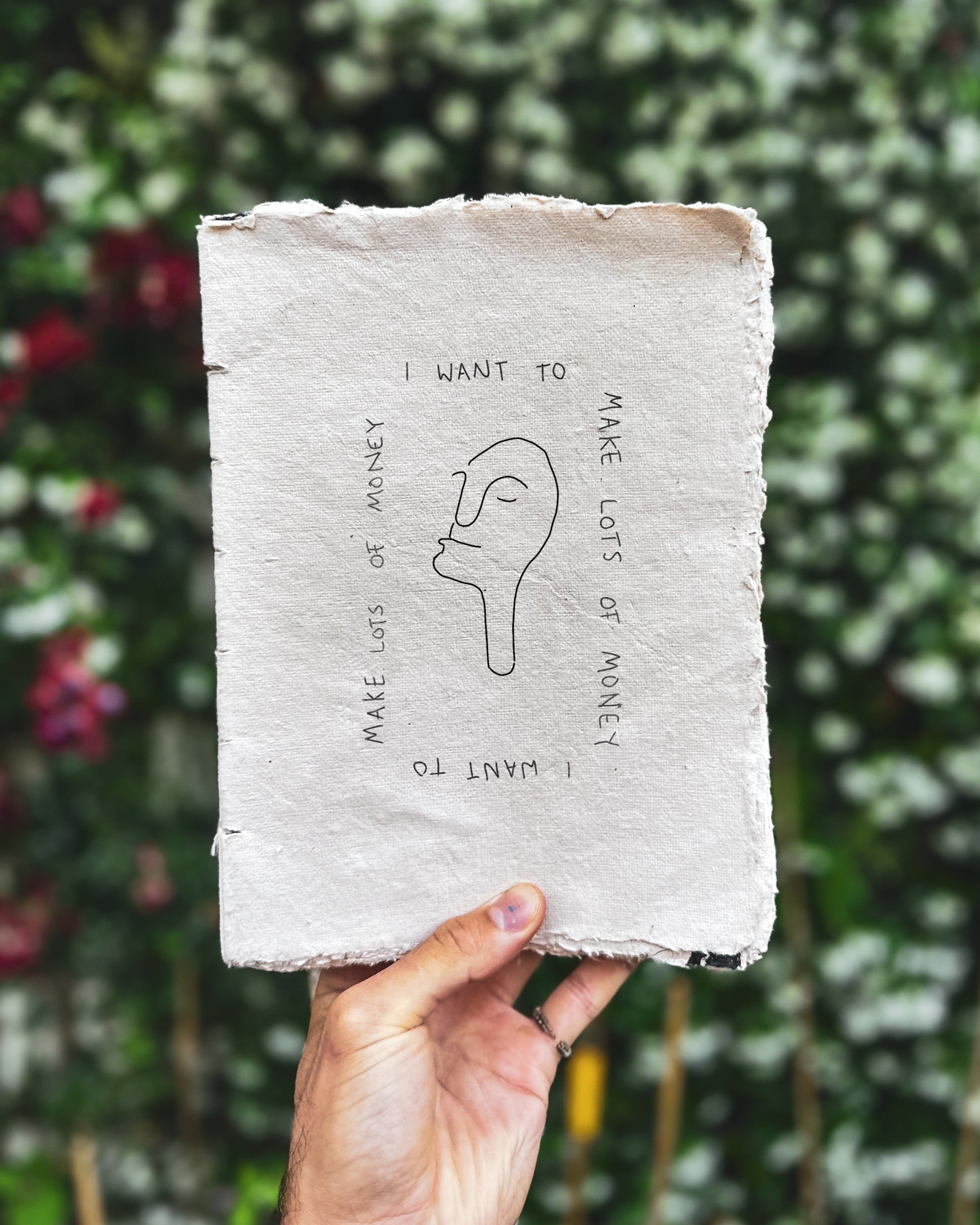 Hand holding a textured notebook with text and a fine line face illustration against a blurred green background. The text says I WANT TO MAKE LOTS OF MONEY in a square 
