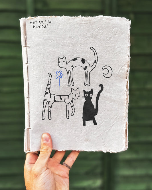 Hand holding a notebook with three sketch-style cat illustrations and text on a blurred green background. Also with a crescent moon and fineline blue flower. Text says WHY AM I SO ANXIOUS?