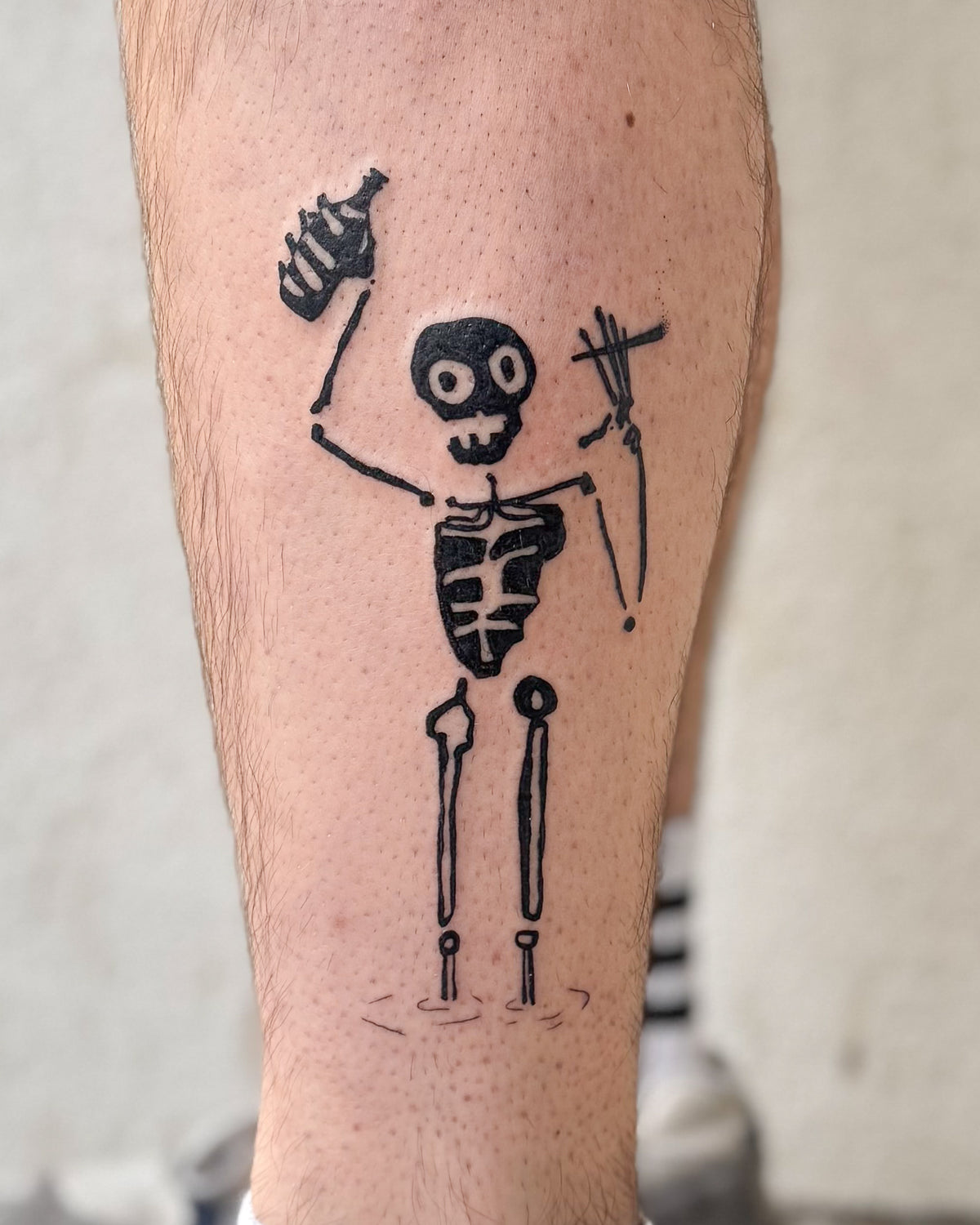 A solid black skeleton tattoo waving hello, on a calf, by Eli Inking