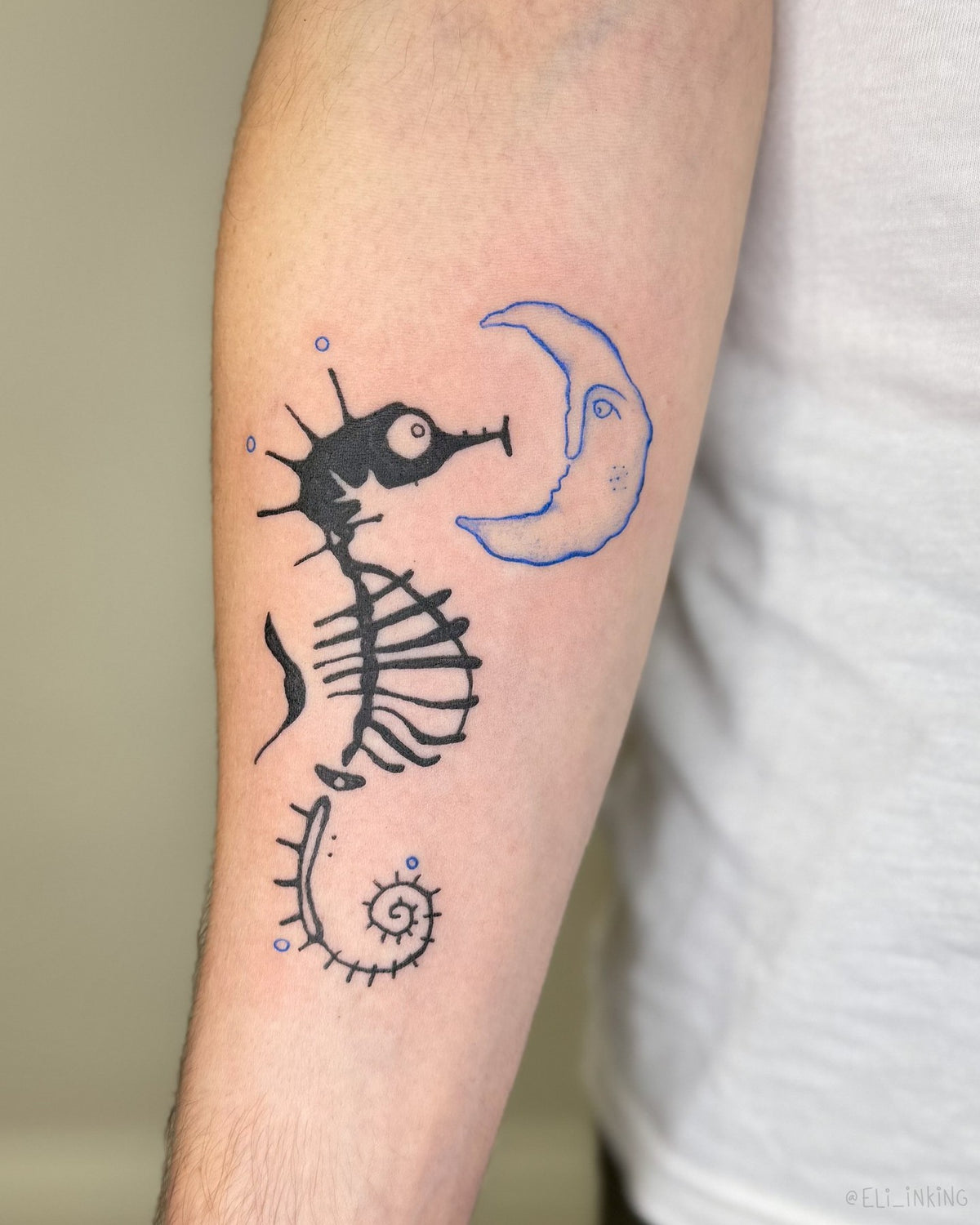 Blue crescent moon face and black skeleton seahorse tattoo on the forearm, by Eli Inking, Bristol