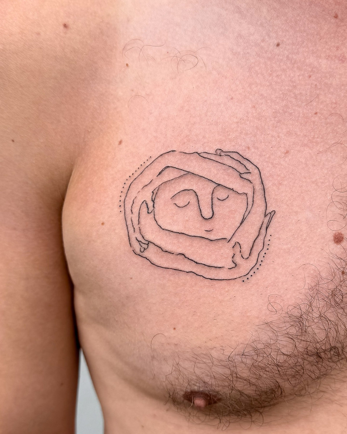 Fine line tattoo of a sleeping face held in a ring of hands, on the chest, by Eli Inking in Bristol