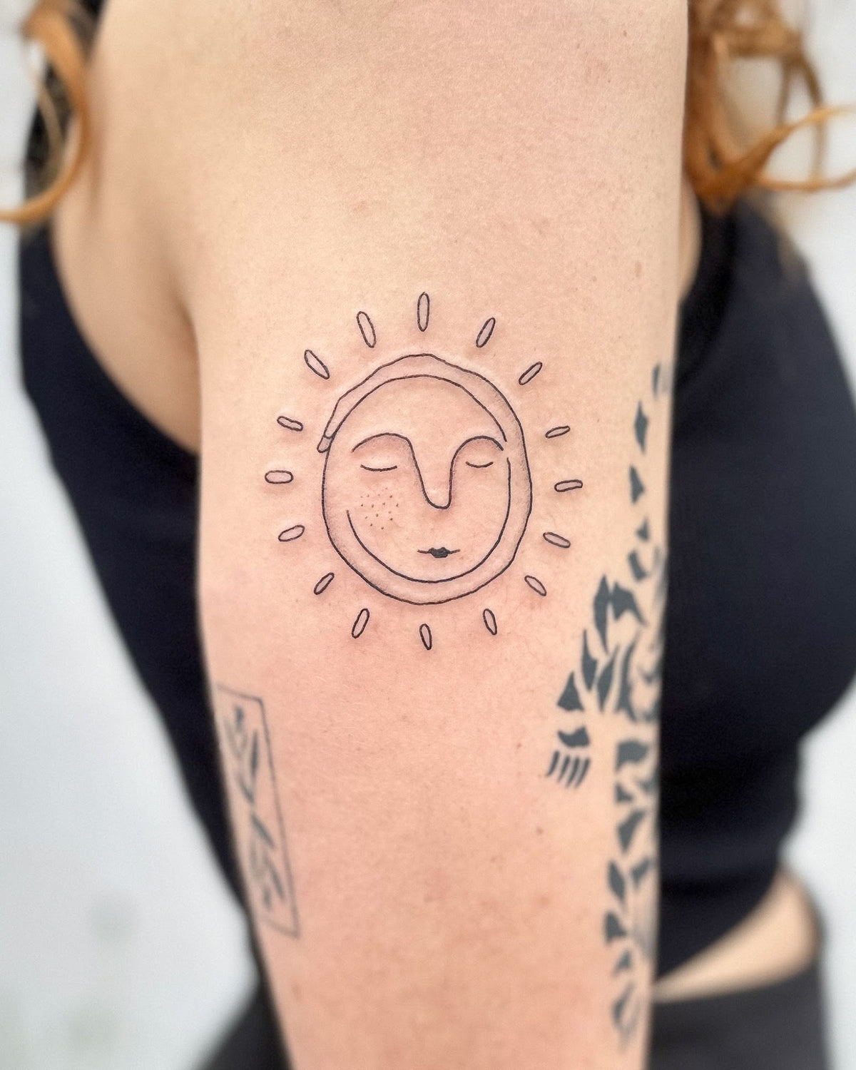 Fine line sun face tattoo with closed eyes and freckles on the back of the arm, by Eli Inking