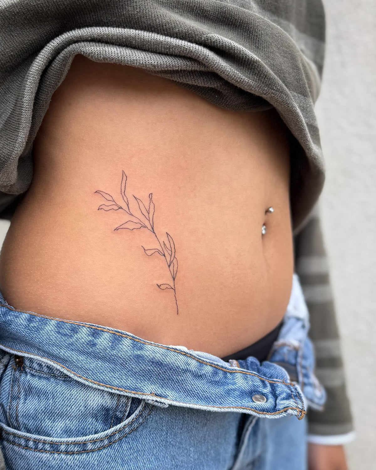 Fine line tattoo of a leafy sprig beside the belly button, by Eli Inking in Bristol