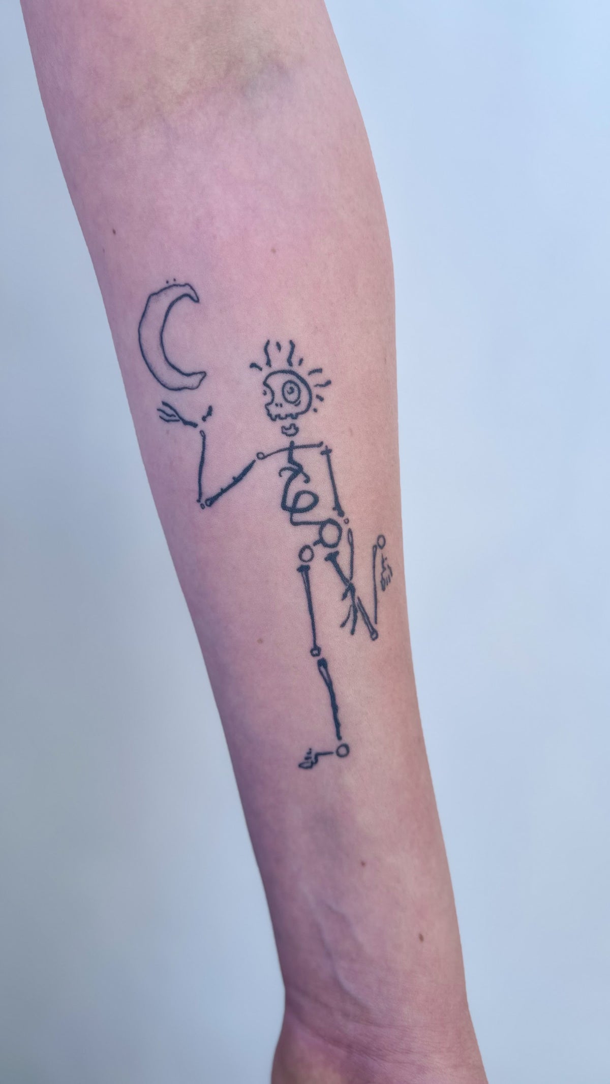 Delicate fine line skeleton tattoo with a sun for a head, holding up a crescent moon, on an inner forearm, by Eli Inking in Bristol