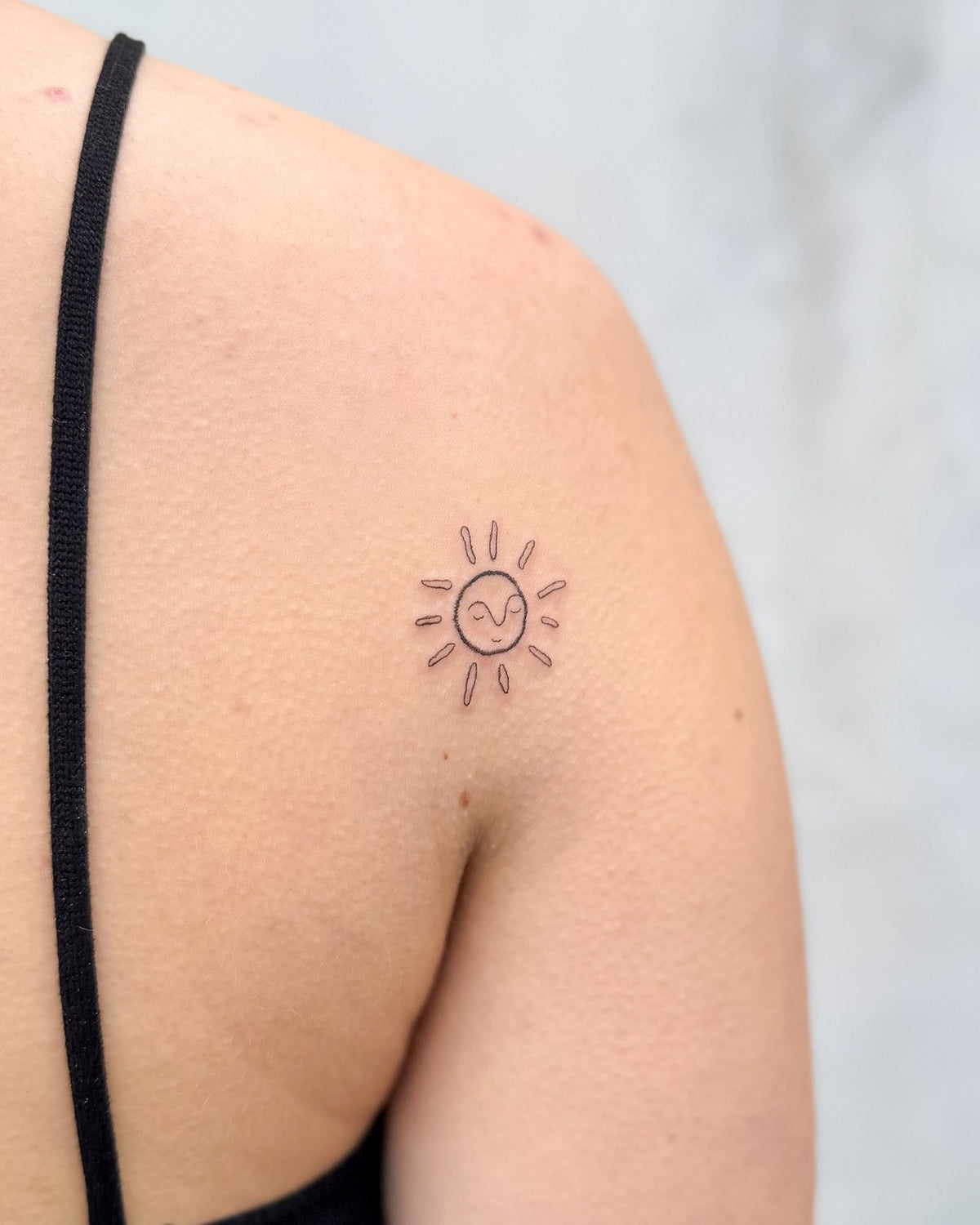 Small fine line sun with a face on the back of the shoulder, by Eli Inking in Bristol