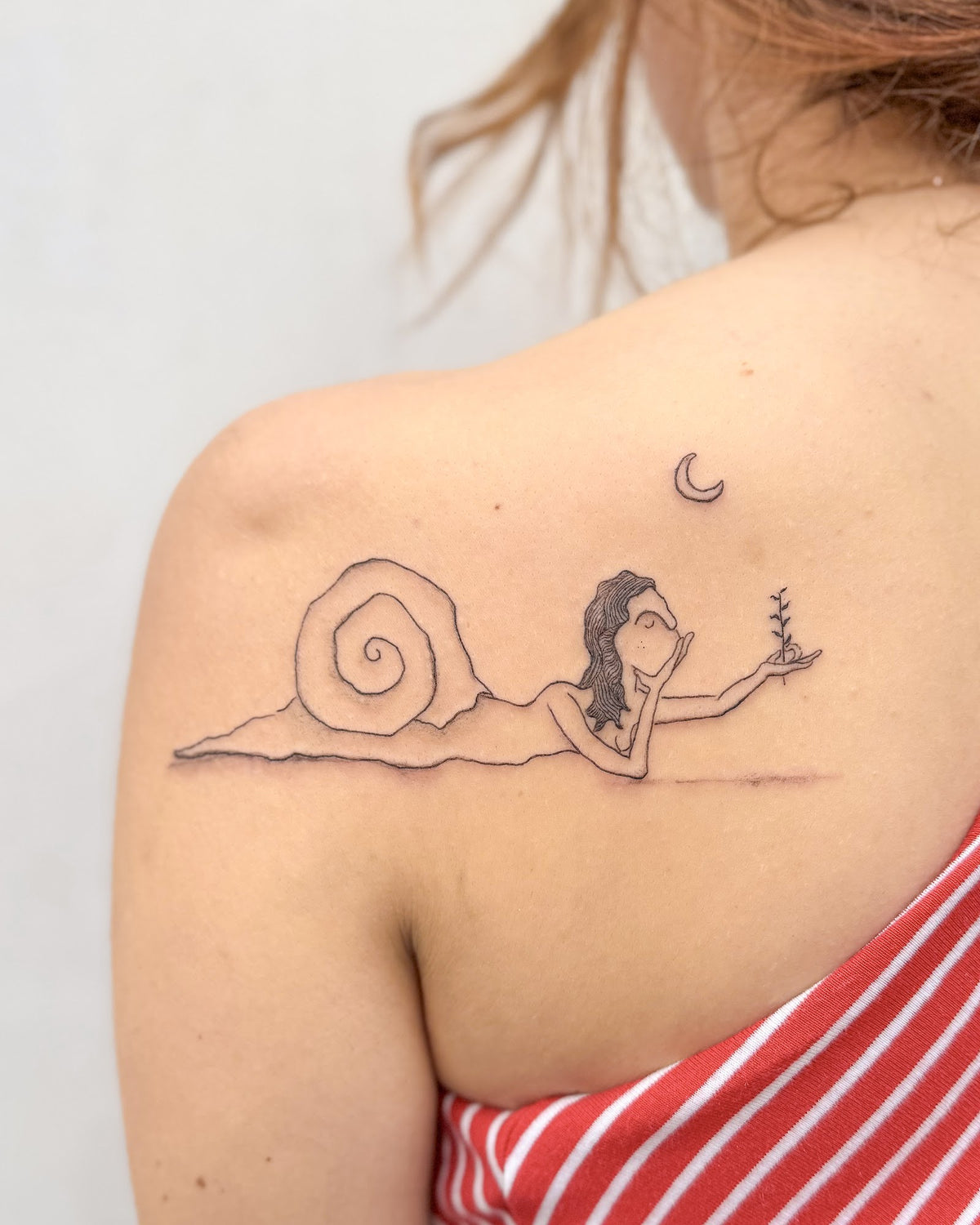Fine line tattoo of a woman with a snail shell holding a sprig under a small moon, on the shoulder, by Eli Inking in Bristol