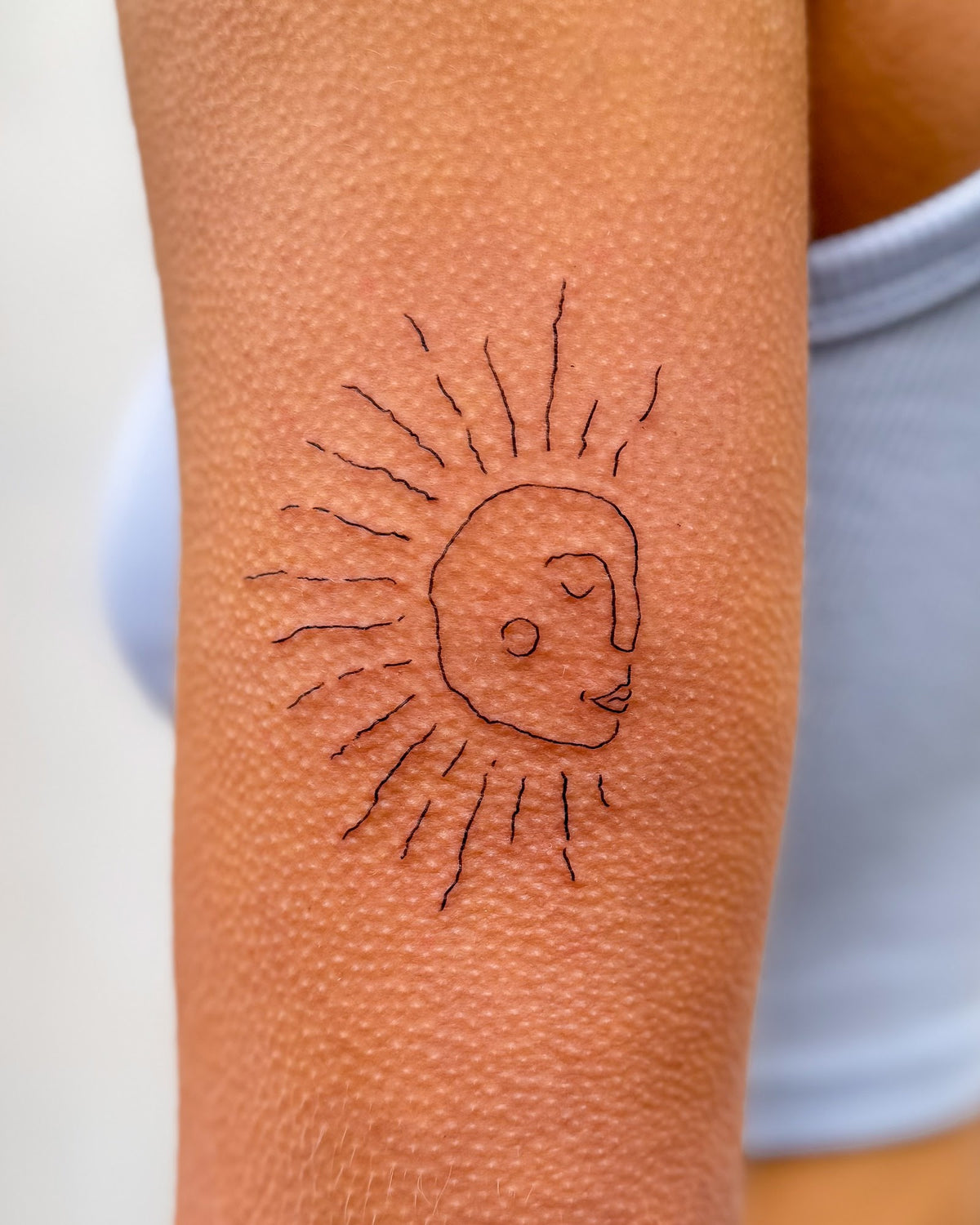 Fine line tattoo of a sun face with spiky rays, on the upper arm, by Eli Inking in Bristol