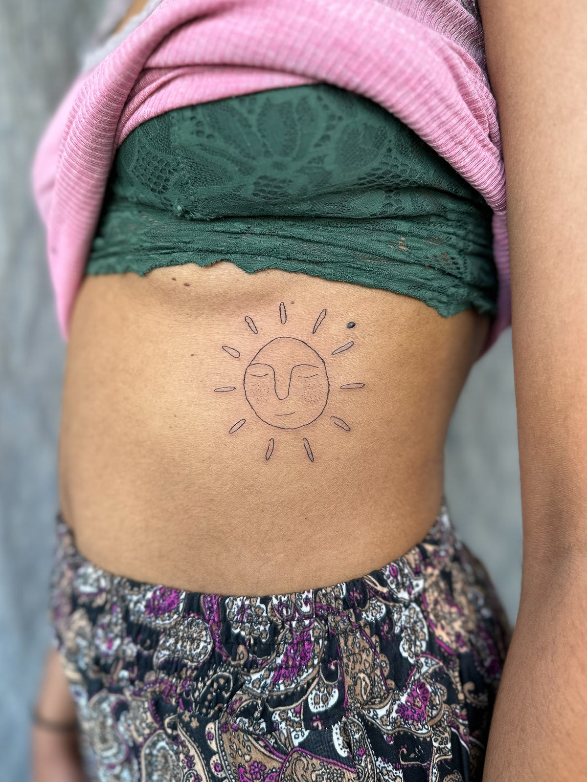 Fine line sun face tattoo with closed eyes on the ribs, by Eli Inking