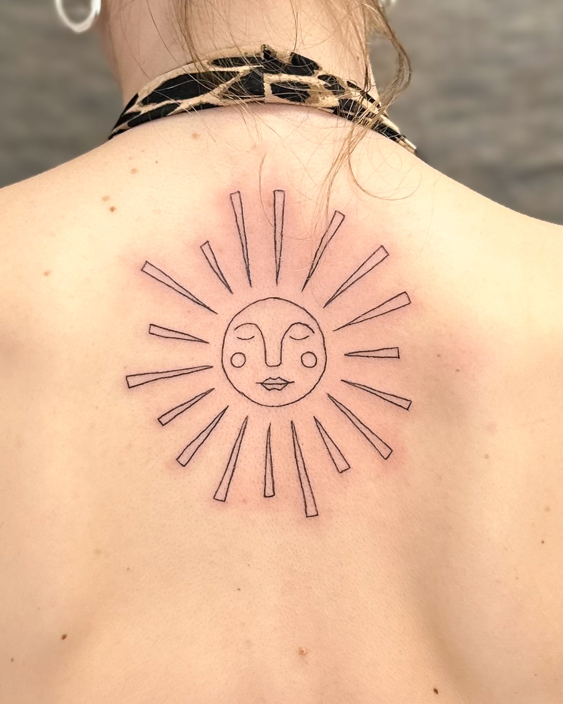 Fine line sun tattoo with a face and long rays on the upper back, by Eli Inking
