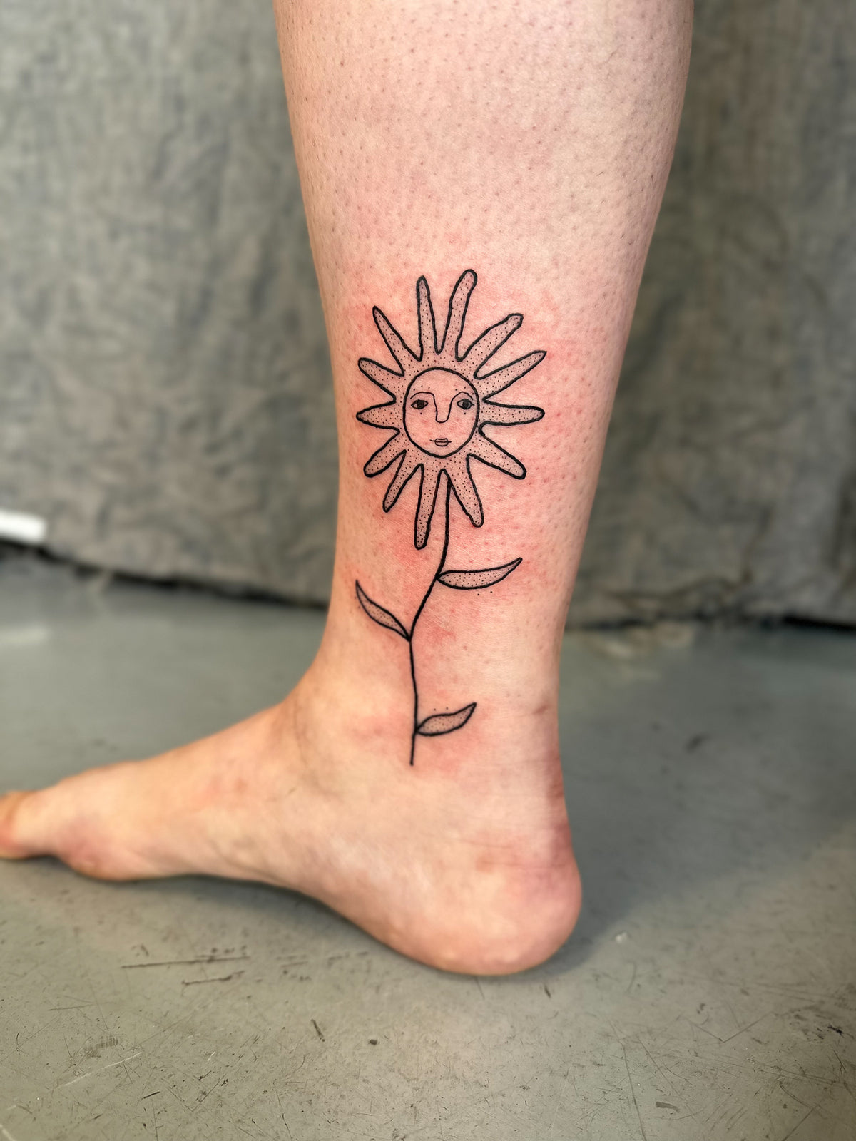 Fine line flower tattoo with a sun face on the ankle, by Eli Inking