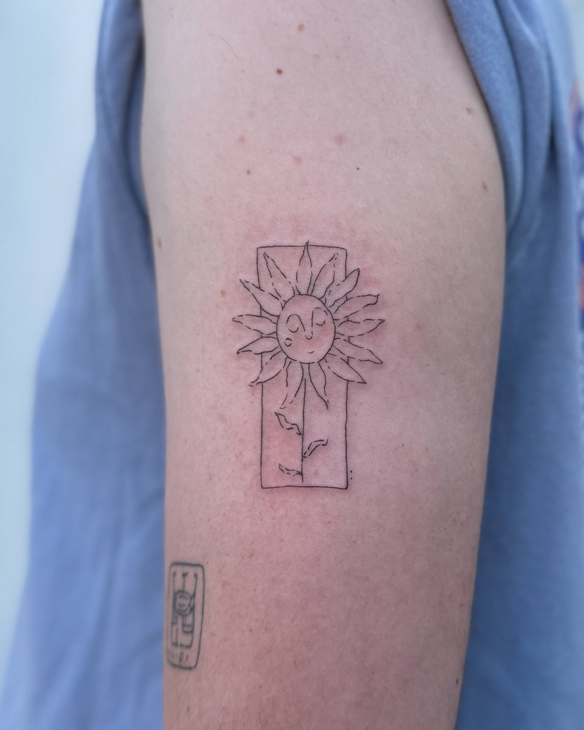 Fine line tattoo of a sunflower with a face inside a tall frame, on the upper arm, by Eli Inking in Bristol