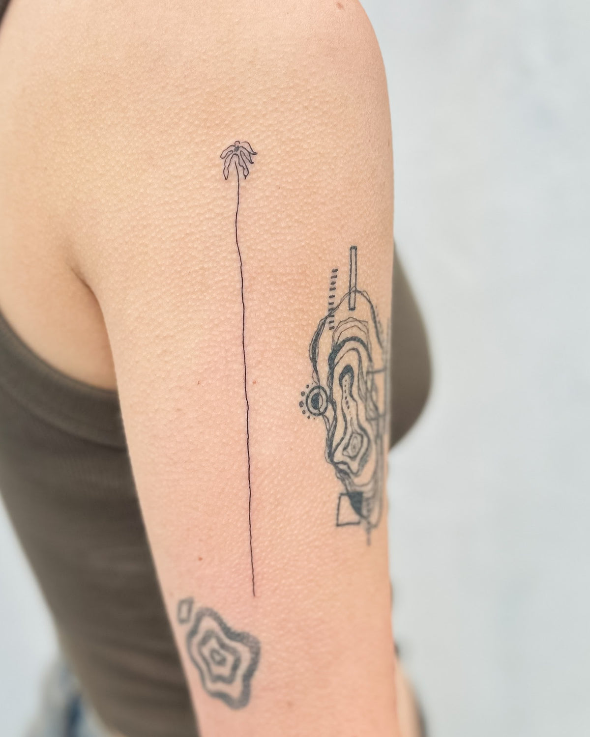 Fine line tattoo of one tall thin flower stem, on the upper arm, by Eli Inking in Bristol