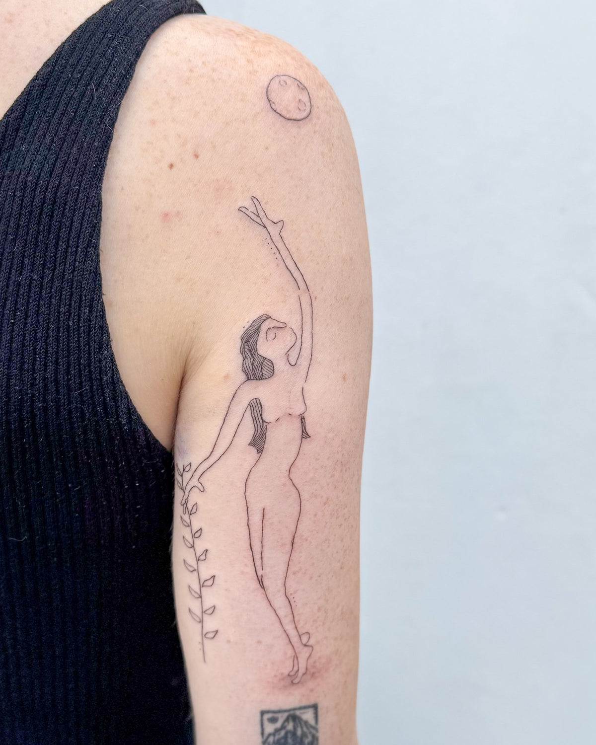 Fine line tattoo of a woman reaching up to a small moon, with a vine, on the upper arm, by Eli Inking in Bristol