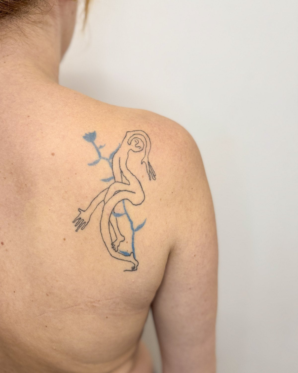 Healed tattoo of a line-drawn figure with a blue flower behind it, on a shoulder