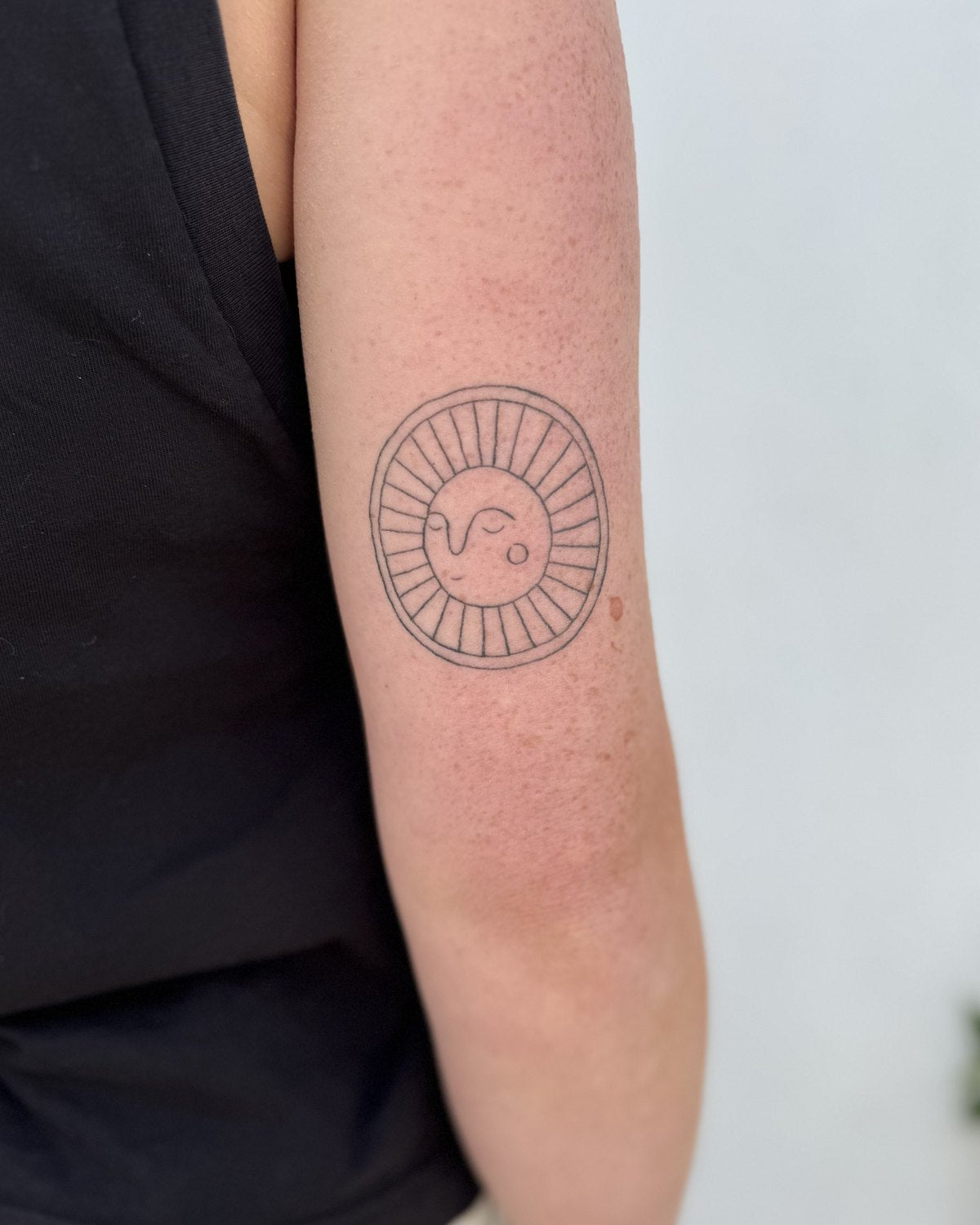 Healed fine line tattoo of a small sun with a face, on an upper arm
