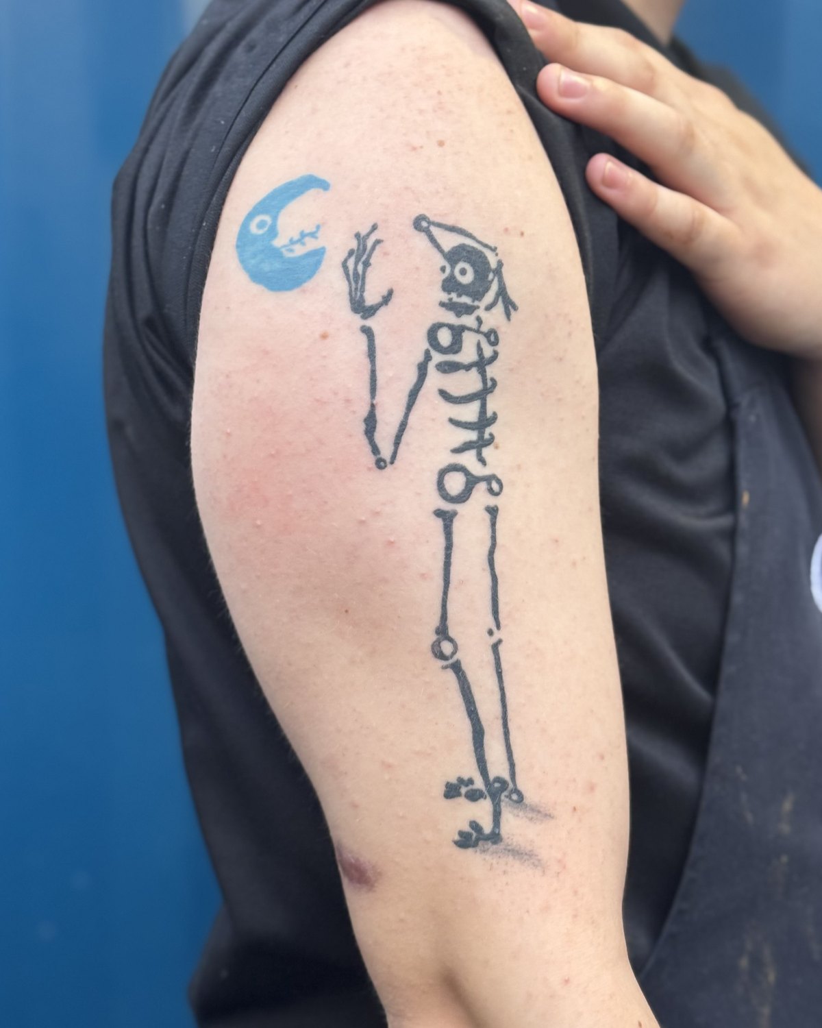 Healed tattoo of a black skeleton figure with a blue-filled crescent moon, on a bicep