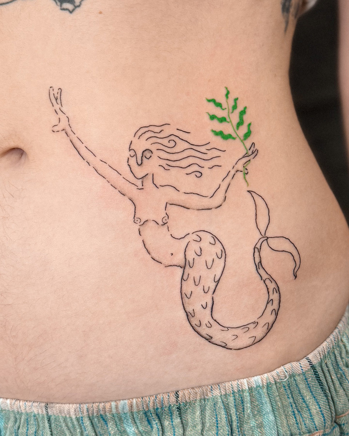 Fine line mermaid holding a green branch, across the stomach, by Eli Inking in Bristol