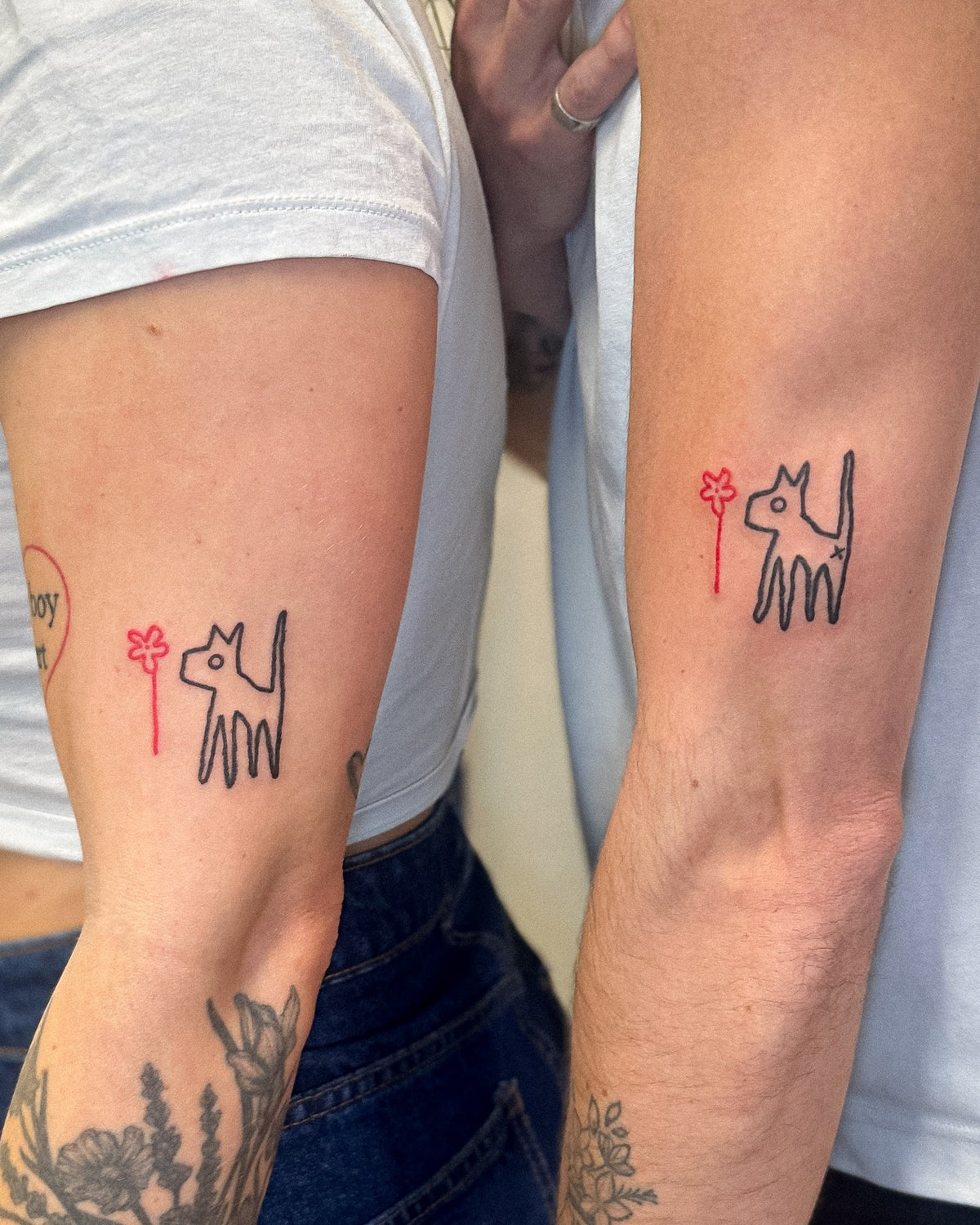 Matching tattoos of a scruffy black dog with a red flower on two people's arms, by Eli Inking in Bristol