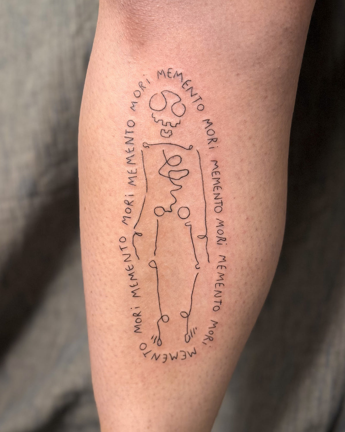 Fine line skeleton tattoo surrounded by an oval of the words memento mori, on a calf, by Eli Inking in Bristol