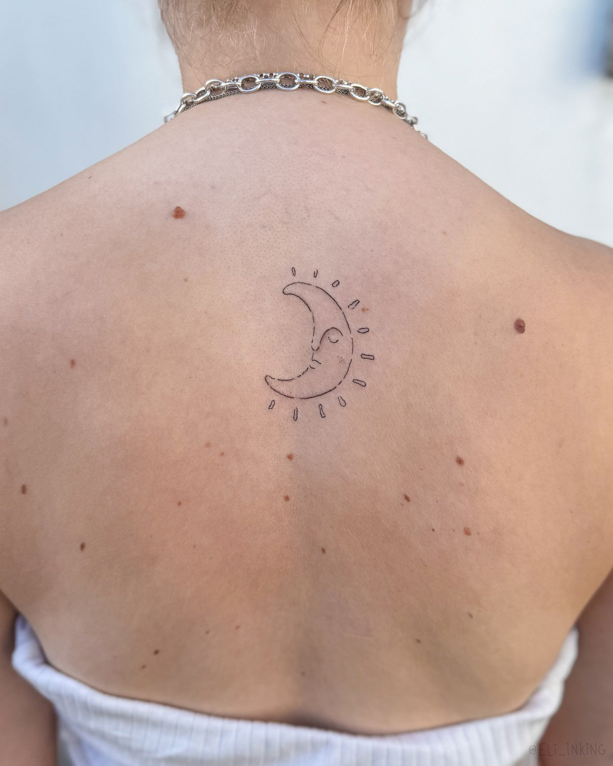 Small fine line crescent moon face tattoo with rays on the upper back, by Eli Inking, Bristol