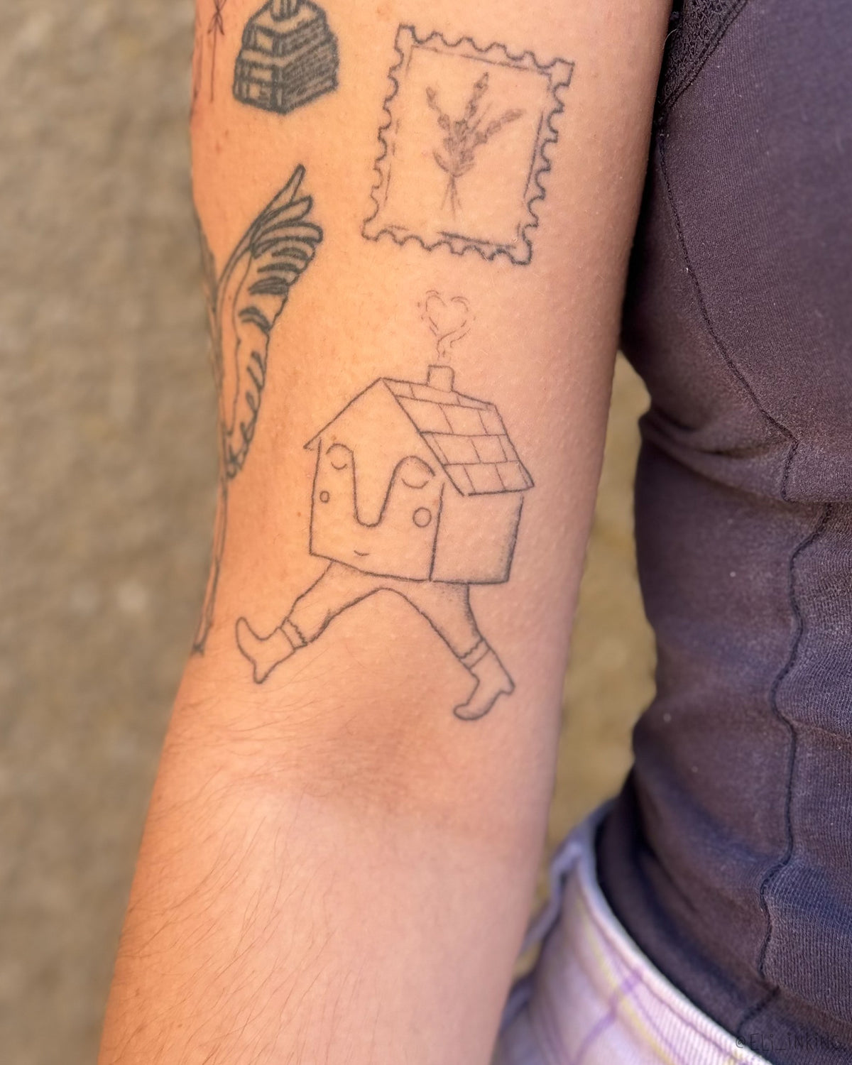 House with legs and a face tattooed on a bicep, placed beside healed work by other artists