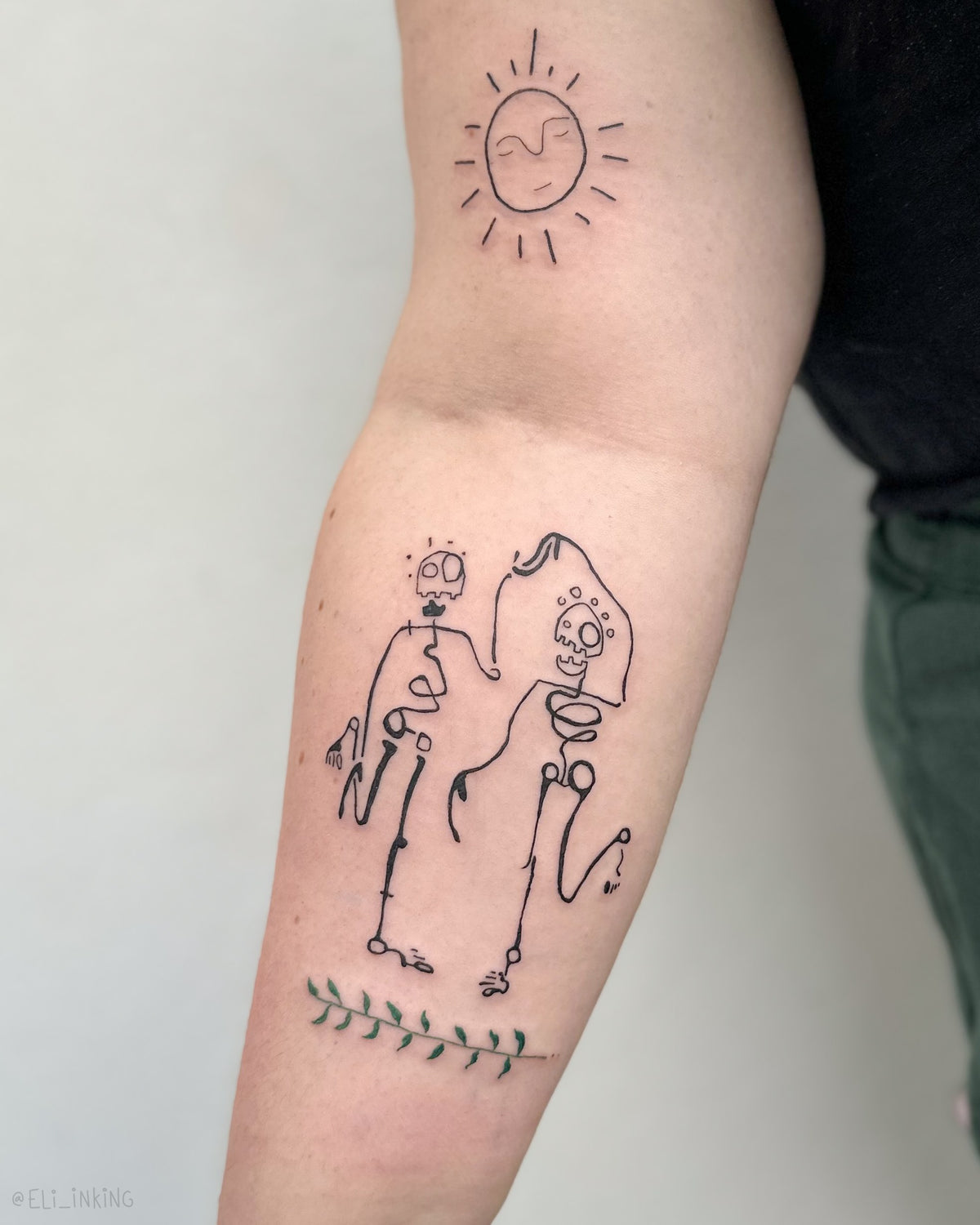 Ornate leaf, skeletons and a sunshine tattooed across an upper arm in a spread-out composition