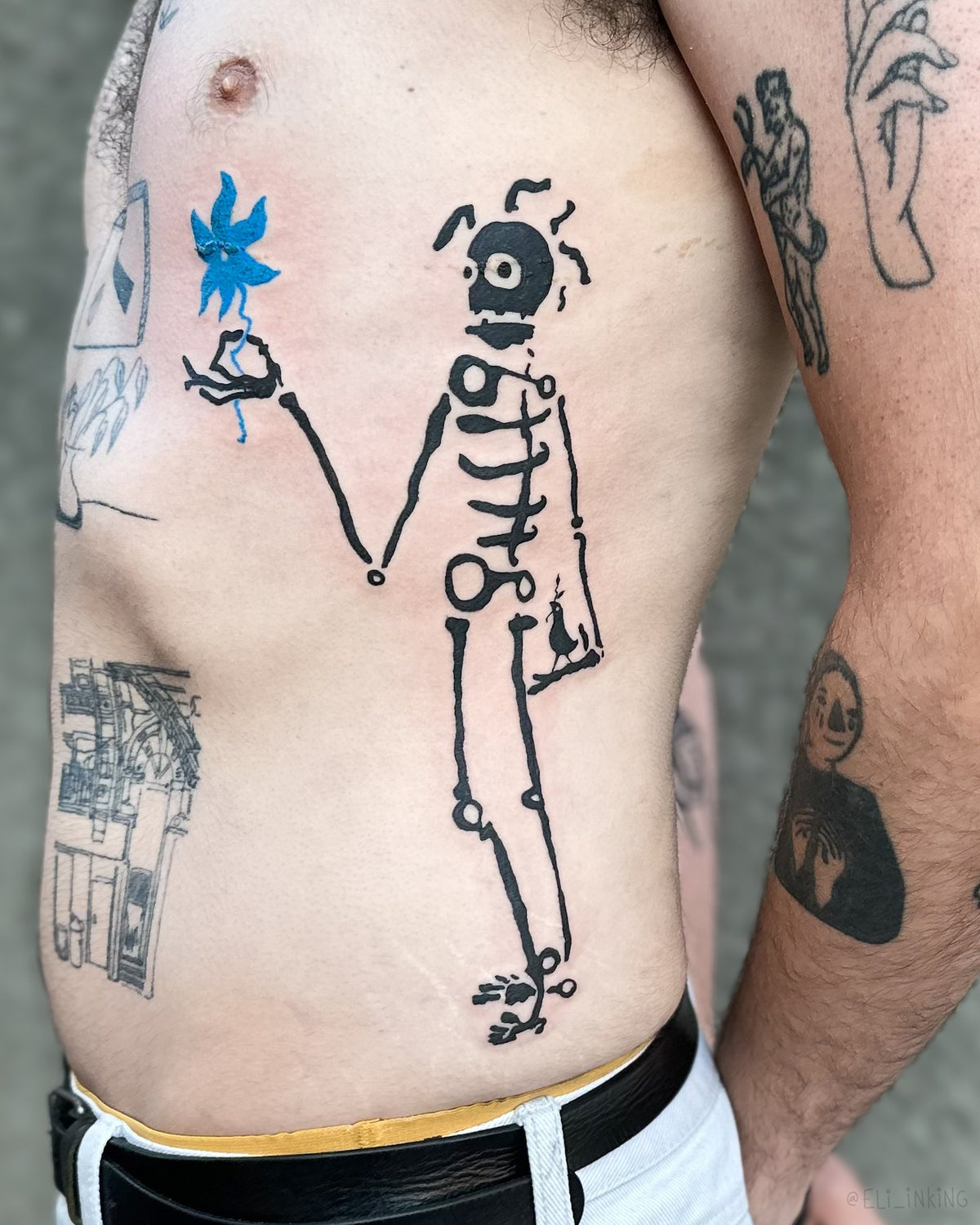 Large blackwork skeleton and flower tattooed across a torso, sitting within a patchwork of existing tattoos