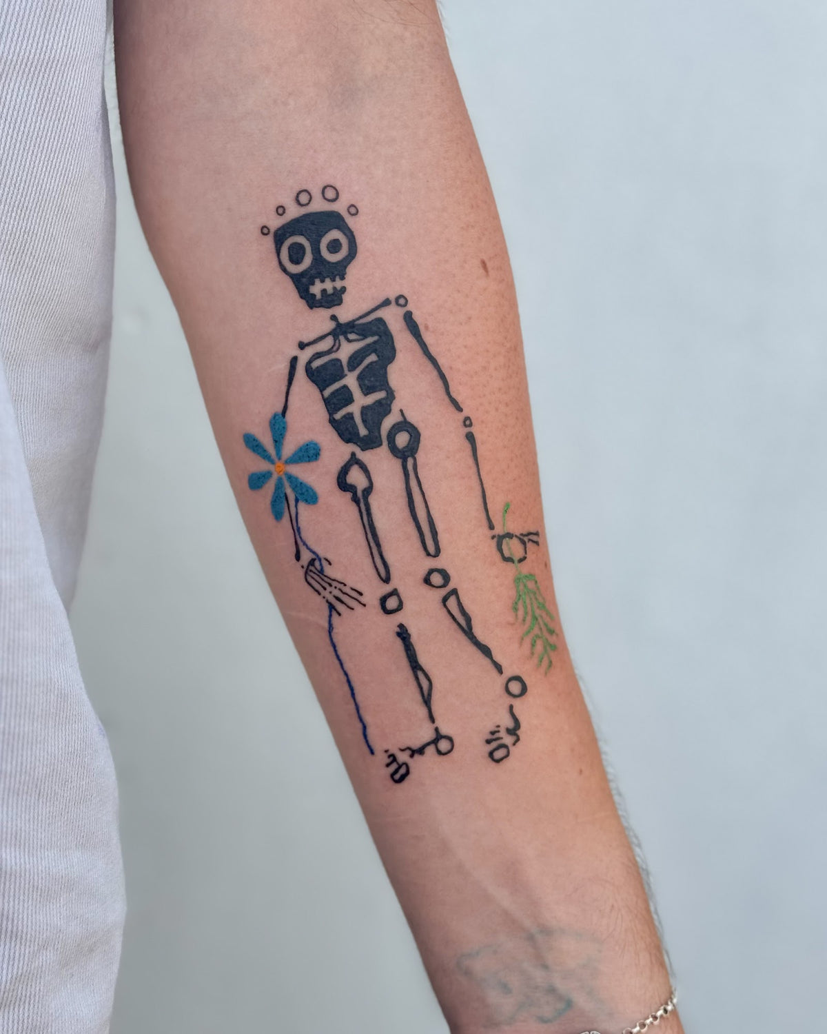 Bold skeleton tattoo holding a blue flower, with a small green sprig, on a forearm, by Eli Inking in Bristol