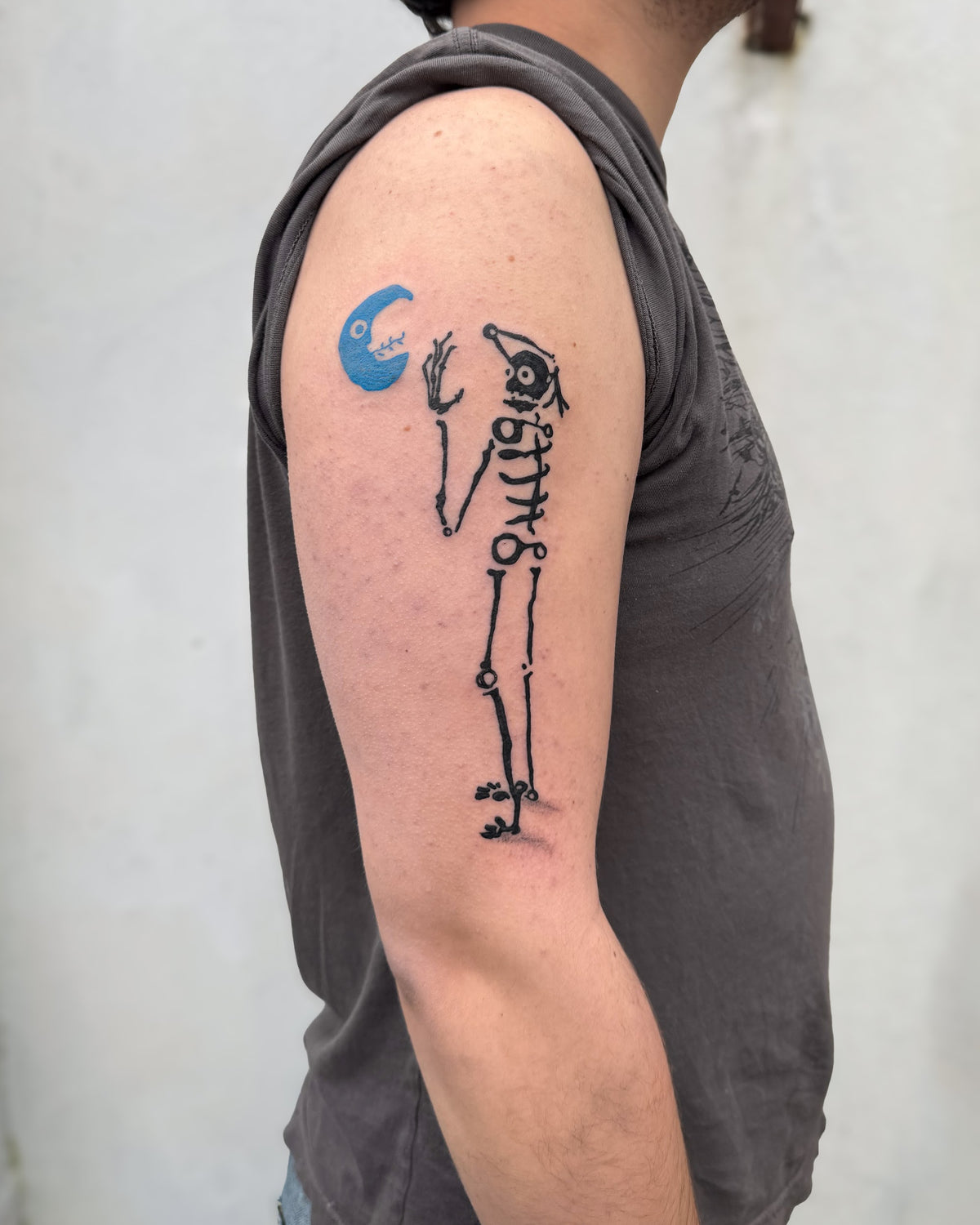 A bold skeleton tattoo face to face with a blue crescent moon, on an upper arm, by Eli Inking