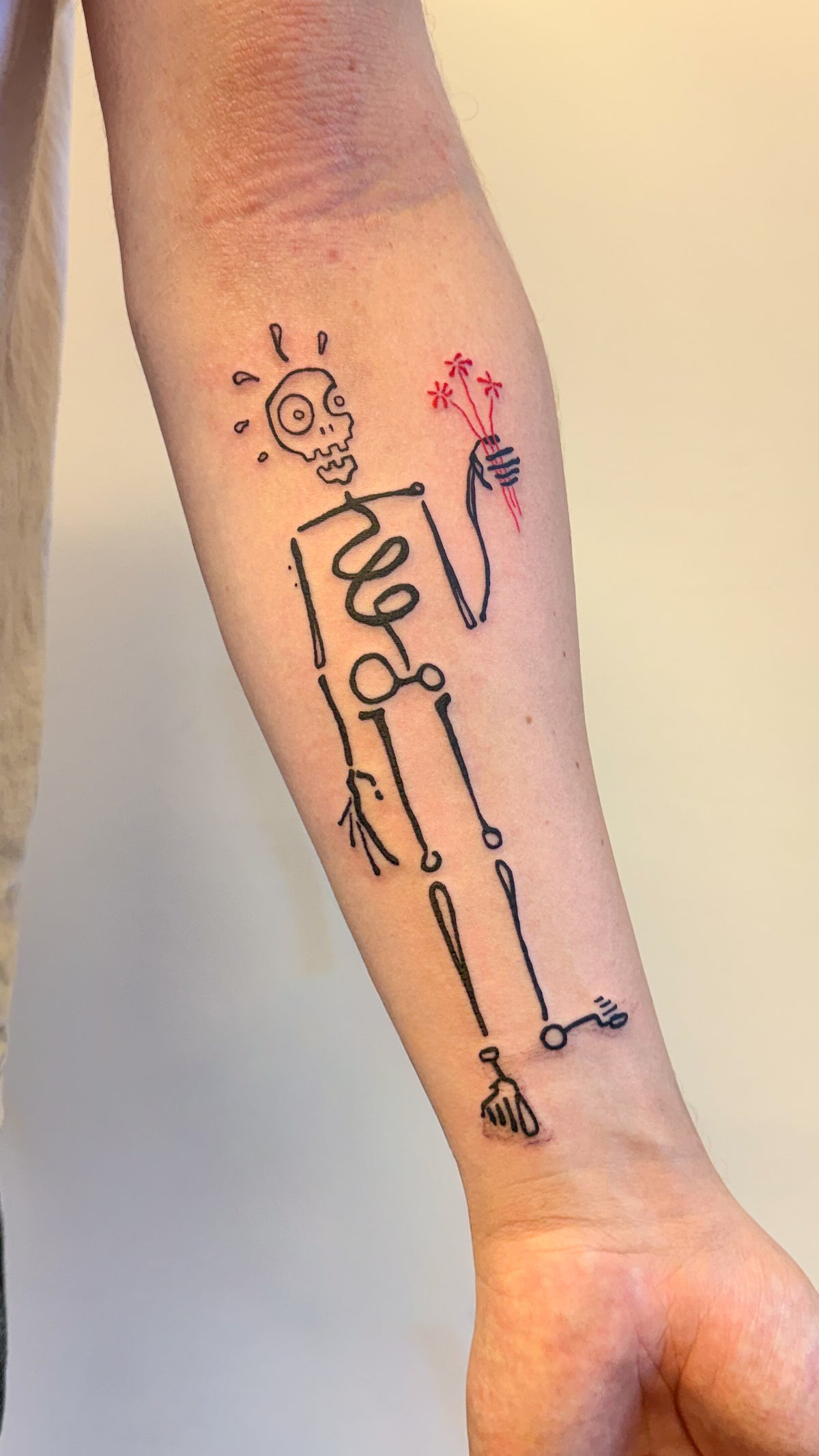 A skeleton tattoo with spiral ribs holding a little bunch of red flowers, on an inner forearm, by Eli Inking