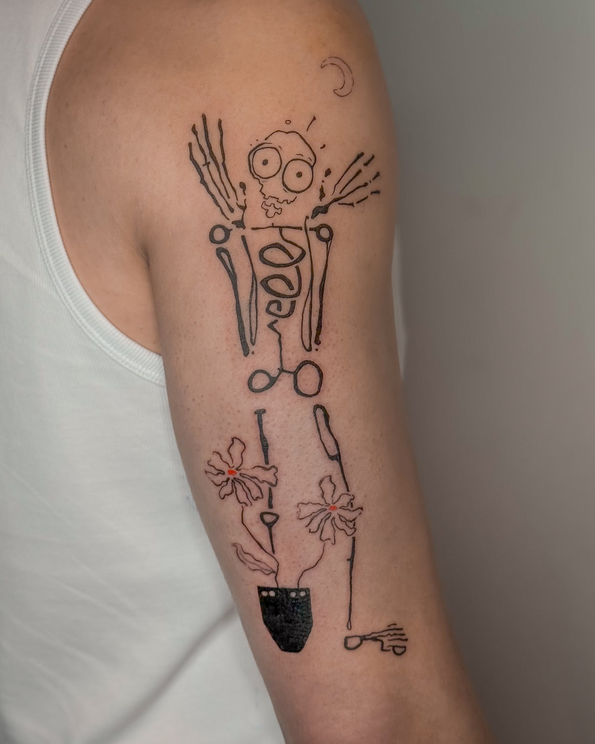 Skeleton tattoo with two red flowers growing from a black pot between its legs, on an upper arm, by Eli Inking in Bristol