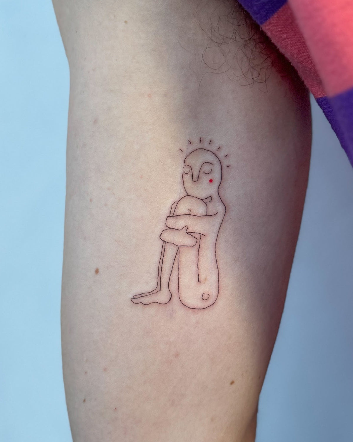 Small red line tattoo of a little figure hugging its knees, on the upper arm, by Eli Inking, Bristol