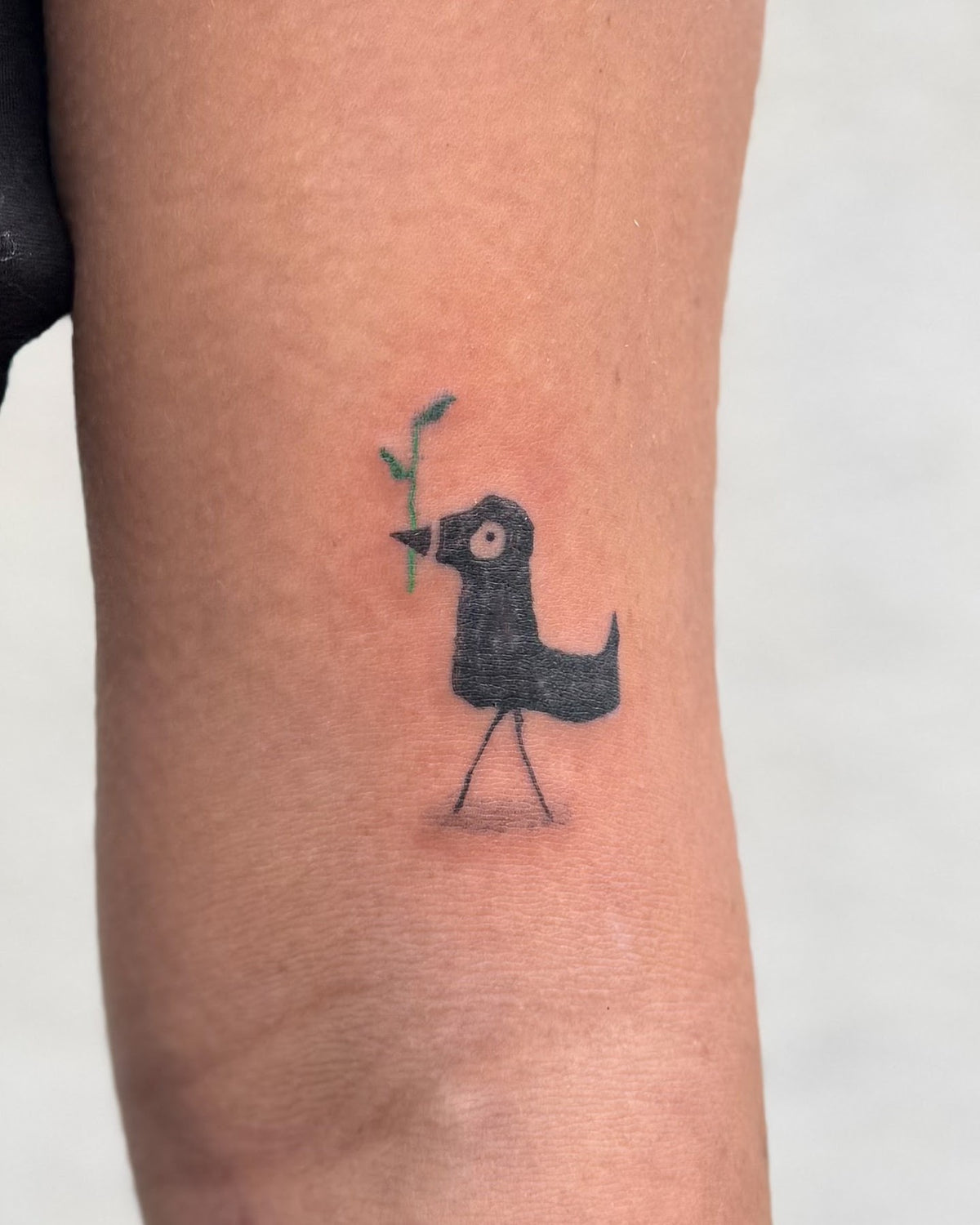 Small bold black bird tattoo holding a green twig, on the upper arm, by Eli Inking, Bristol