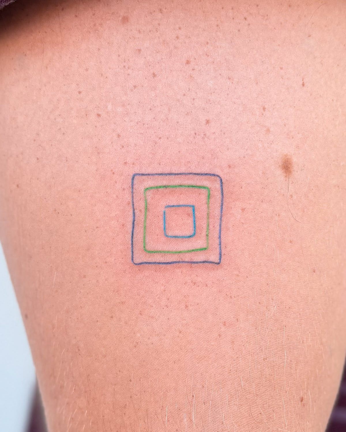 Small tattoo of nested squares in blue, green and grey lines, on the upper arm, by Eli Inking, Bristol