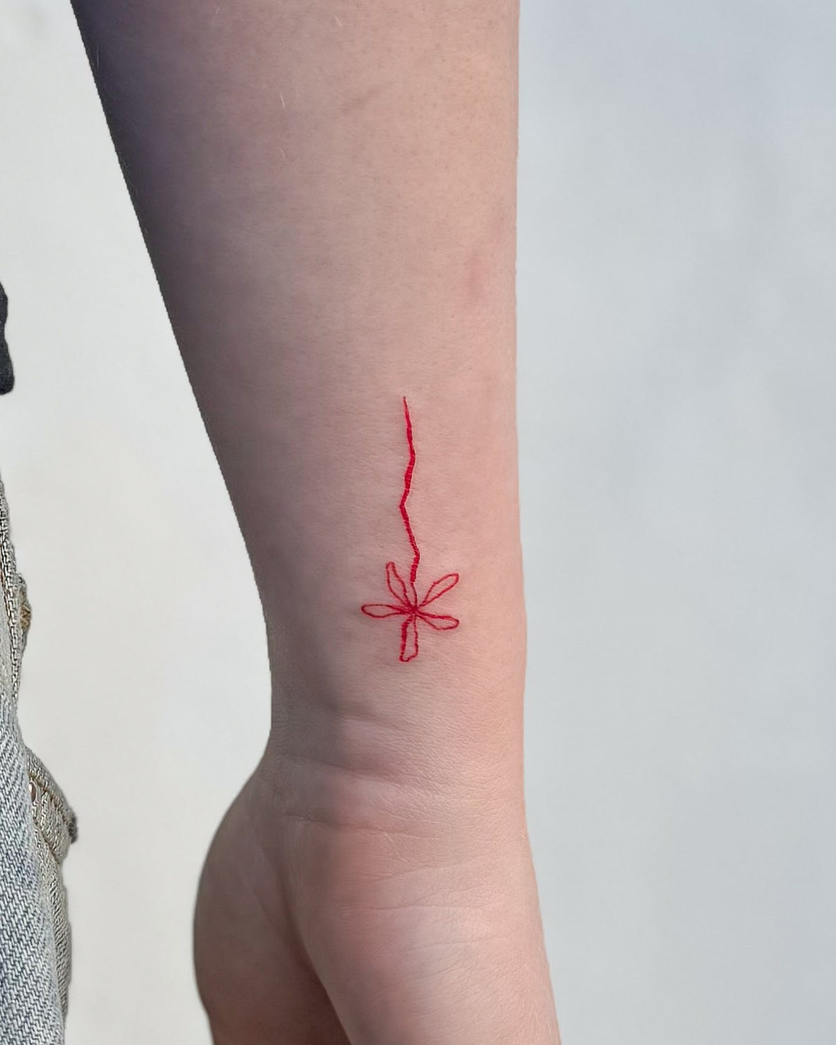 Small fine line tattoo of a red star-shaped flower with a wavy stem, on the wrist, by Eli Inking, Bristol