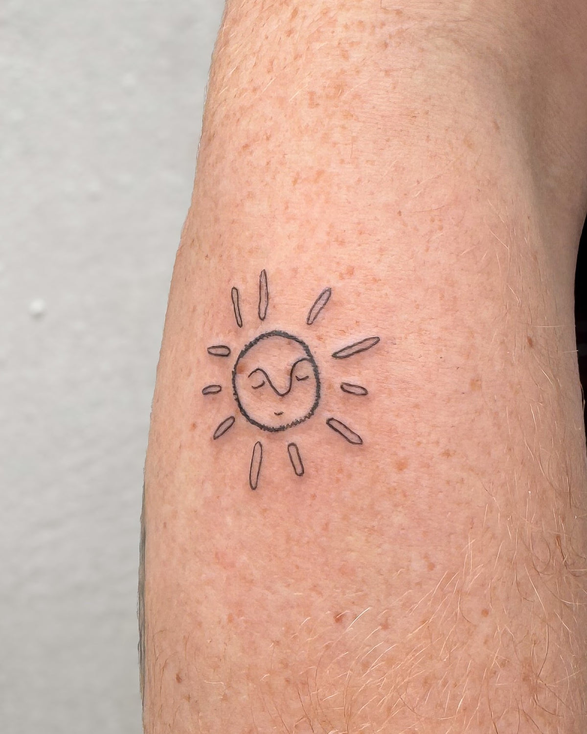 Small fine line smiling sun tattoo on the forearm, by Eli Inking, Bristol