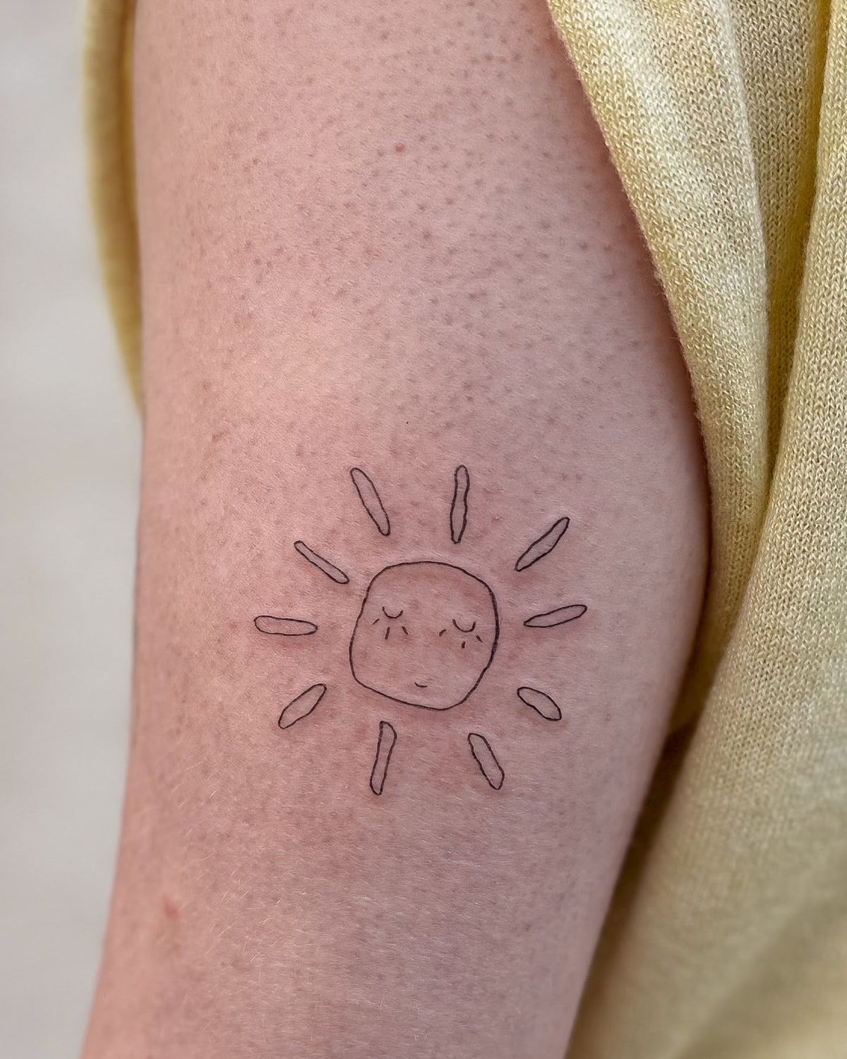 Small fine line sun tattoo with a sleepy face, on the upper arm, by Eli Inking, Bristol