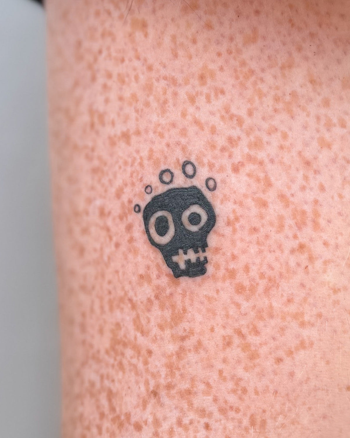 Tiny bold black skull tattoo with a halo of dots, on the upper arm, by Eli Inking, Bristol