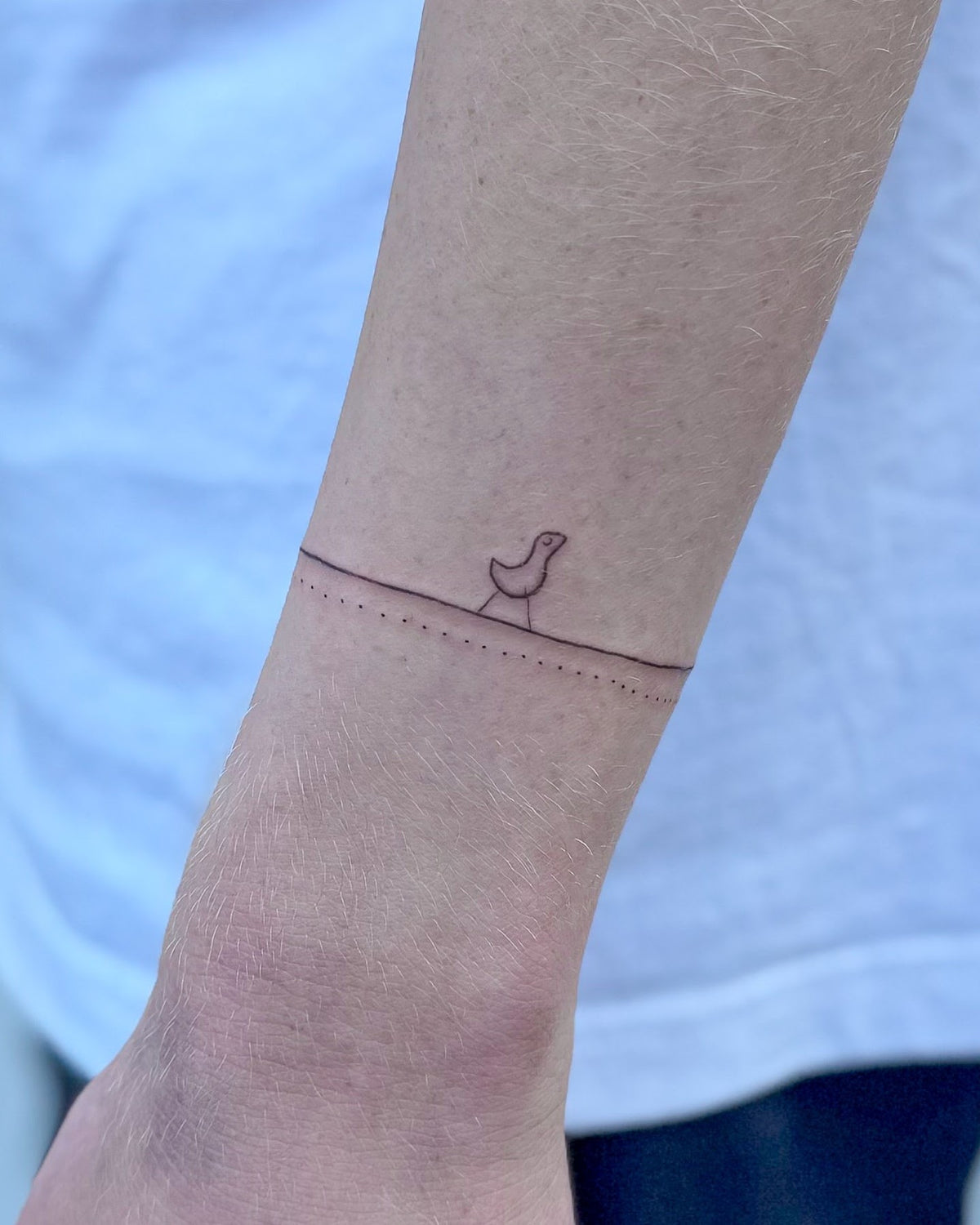 Fine line tattoo of a thin line round the wrist with a tiny bird sitting on it, by Eli Inking, Bristol