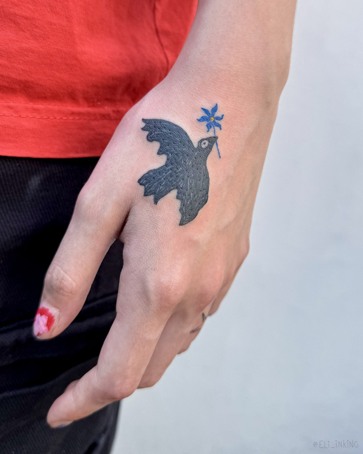 Small black bird tattoo holding a blue flower, on the back of a hand, by Eli Inking in Bristol