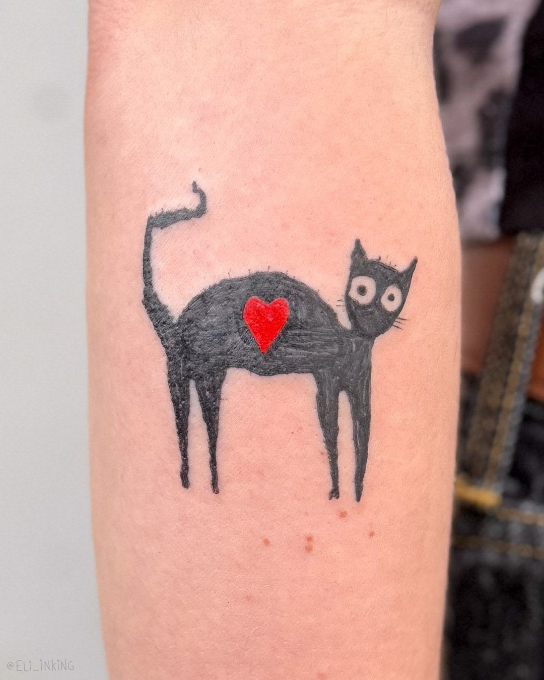 Cute black cat tattoo with a small red heart on its side, on a forearm, by Eli Inking in Bristol