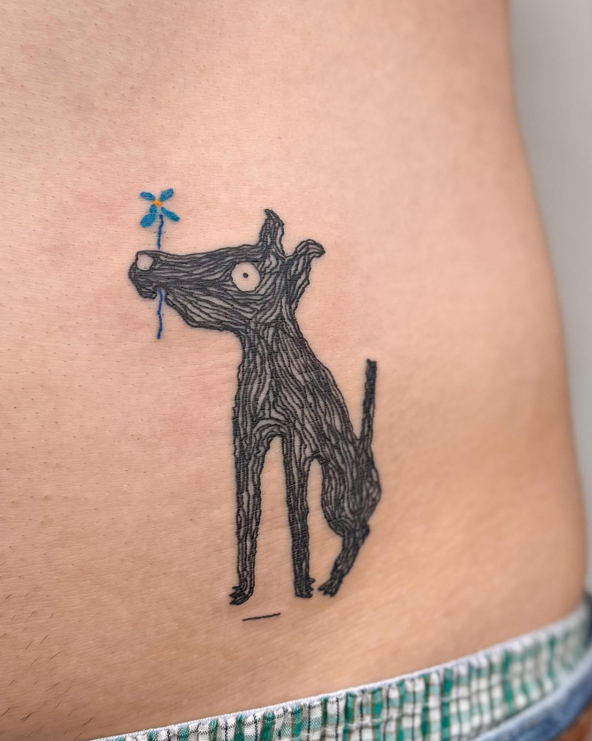 Bold black dog tattoo holding a small blue flower in its mouth, on a hip, by Eli Inking in Bristol