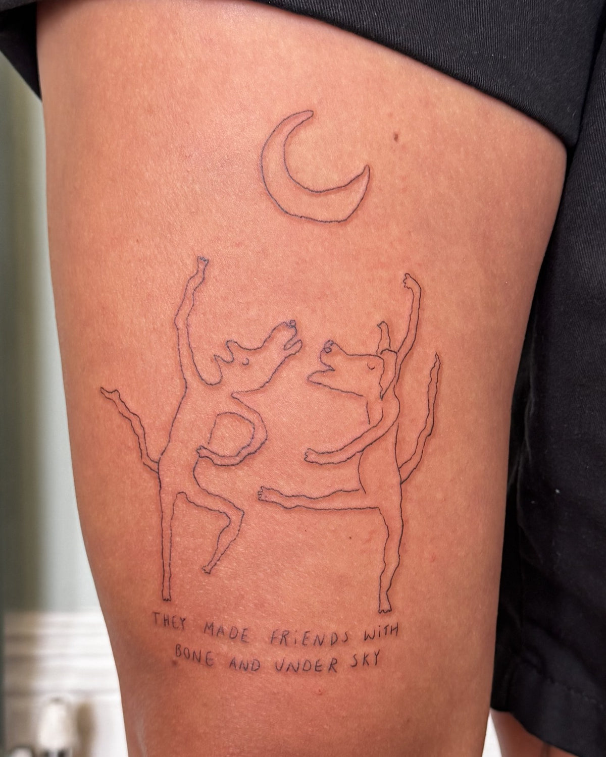 Fine line tattoo of two dogs dancing together under a crescent moon, with a hand-lettered line beneath, on a thigh, by Eli Inking in Bristol