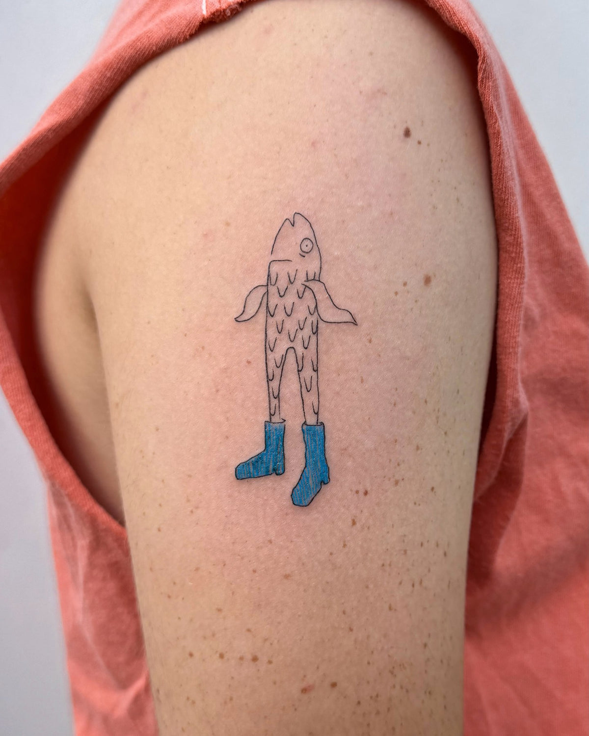 Whimsical fine line tattoo of a fish standing in a pair of blue boots, on an upper arm, by Eli Inking in Bristol