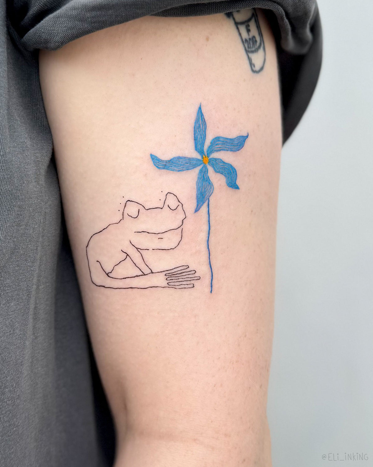 Line drawn frog tattoo sitting beside a tall blue flower, on an upper arm, by Eli Inking in Bristol