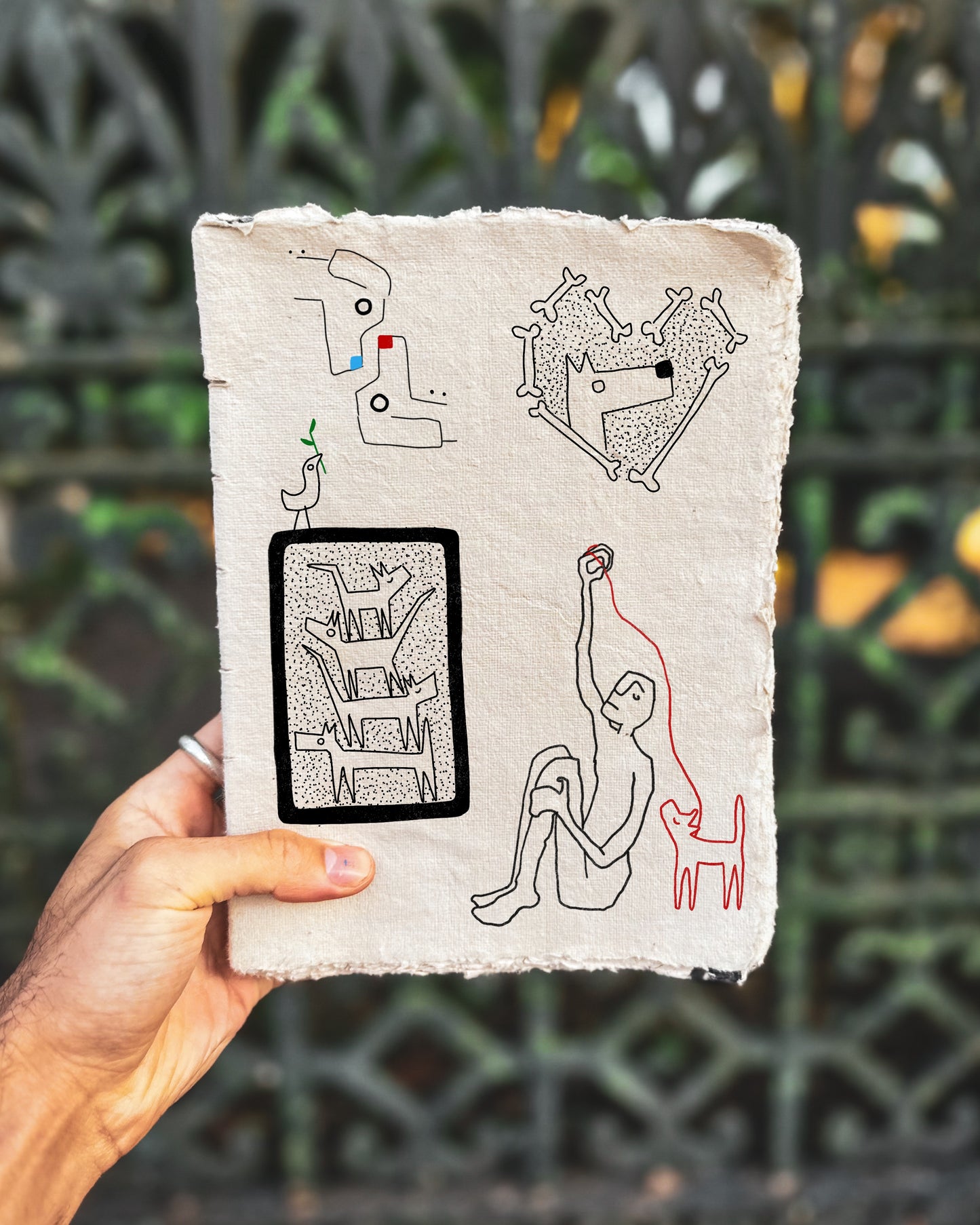 Hand holding a small notebook with black and red fine line drawings of dogs against a blurred green background