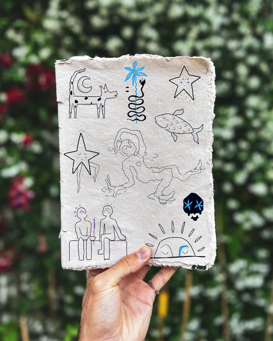Hand holding a piece of paper with drawings against a blurred green background. The fineline drawings include a person floating in water, a star with a face, a rising sun with a face and a fish.