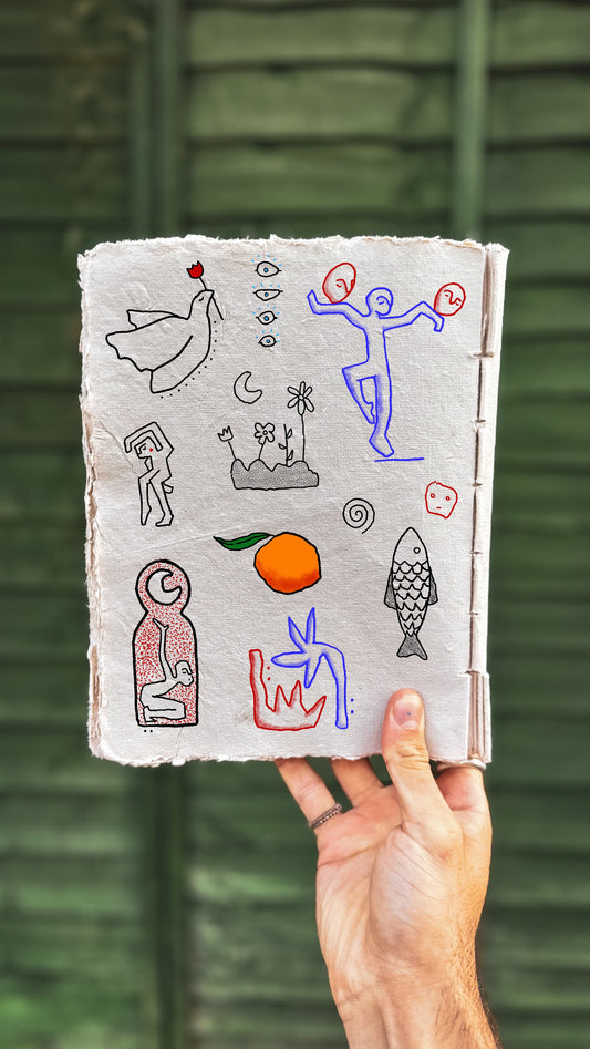 Hand holding a notebook with colorful drawings against a green wooden background. The drawings include minimalist eyes, fineline sprials and textural shading. Also a fish, dove and figures dancing. 